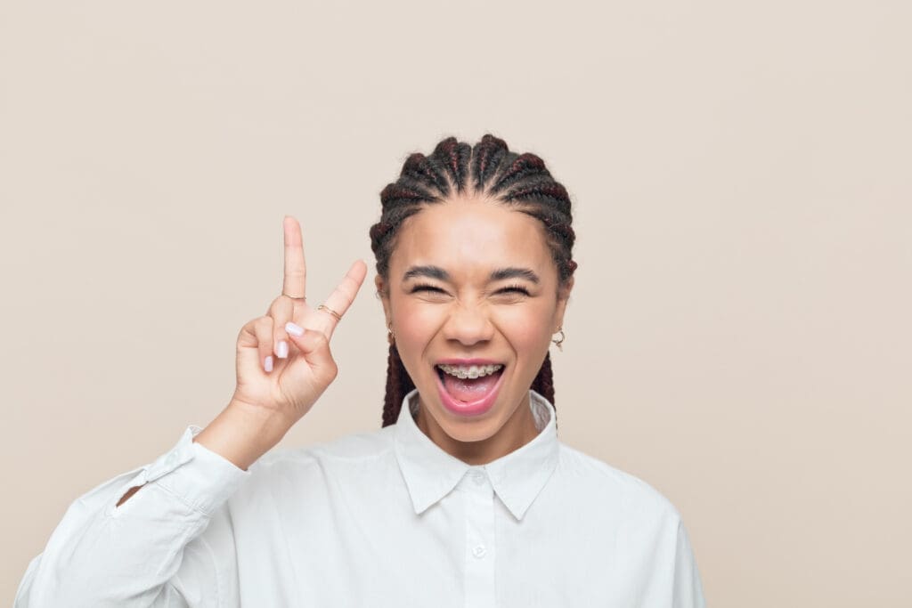 Playful multiracial teenage girl with braided hair wearing white shirt showing peace sign and laughing at camera . Studio shot, beige background.