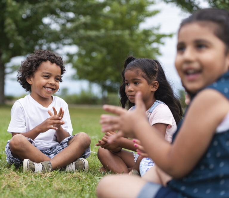 An adorable group of multi-ethnic children are outside sitting in the grass one sunny afternoon. They are clapping and smiling while sitting cross-legged and singing a song.