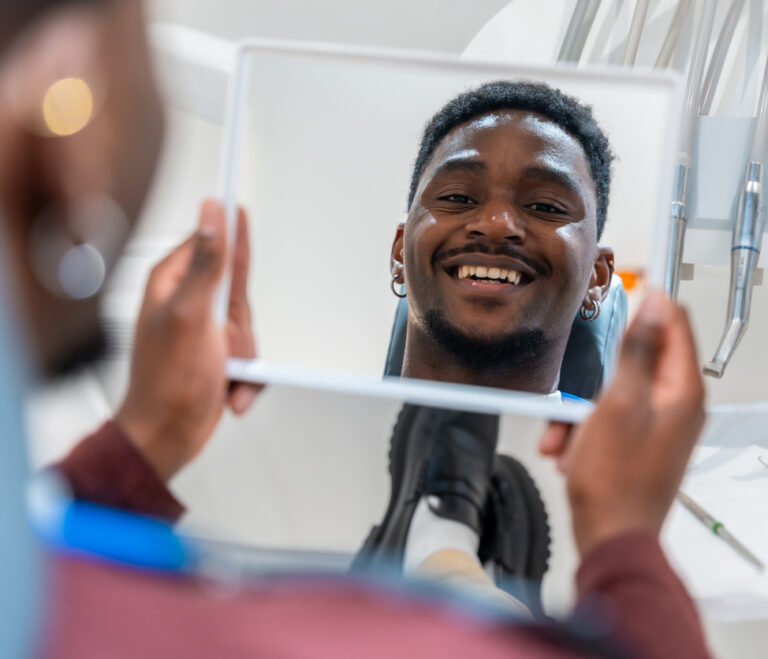 Young man checks his teeth in the mirror at the dental clinic, happy with the results of the treatment